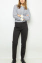 A person stands against a white wall wearing a striped button-up shirt, a pantalon droit 7/8 in matière stretch, and black shoes, with arms crossed and face partially obscured by hair.