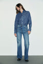 A person stands against a plain background wearing a denim brut button-up shirt tucked into high-waisted jean flare blue jeans and black shoes.