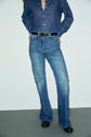 A person wearing a blue denim brut shirt tucked into high-waisted jean flare (taille haute) jeans with a black belt and black shoes stands against a plain light-colored background.