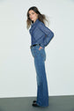 A woman stands sideways against a plain background, wearing a long-sleeve denim shirt tucked into high-waisted, jean flare blue jeans with a black belt and black shoes. Her hair is loose and she has a neutral expression.