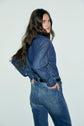 A person with long brown hair, wearing a denim brut shirt and jean flare taille haute jeans, stands sideways with hands in back pockets, looking over their shoulder against a plain light background.