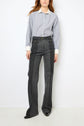 A person wearing a blue striped button-up shirt with white collar and cuffs, paired with a taille haute jean flare in dark gray denim bicolore and black shoes, standing against a plain white wall.