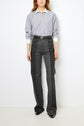 A person stands against a white wall wearing a blue and white striped button-up shirt and high-waisted denim bicolore jeans with front pockets and black shoes. The jean flare silhouette adds a touch of retro style. Their face is not visible.