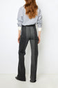 A woman stands facing away, wearing a blue long-sleeve shirt and high-waisted, dark gray jeans with jean flare legs, in front of a plain white background.