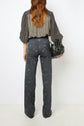 Woman with long, wavy hair seen from behind, wearing a black-and-white checkered blouse and patterned jean flare taille haute, holding a black handbag. She stands against a plain white background.