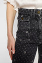Close-up of a person wearing black taille haute jeans with a small motif à pois, front pockets with flaps and metallic buttons, and a white short-sleeved blouse. Only the torso and one arm are visible.