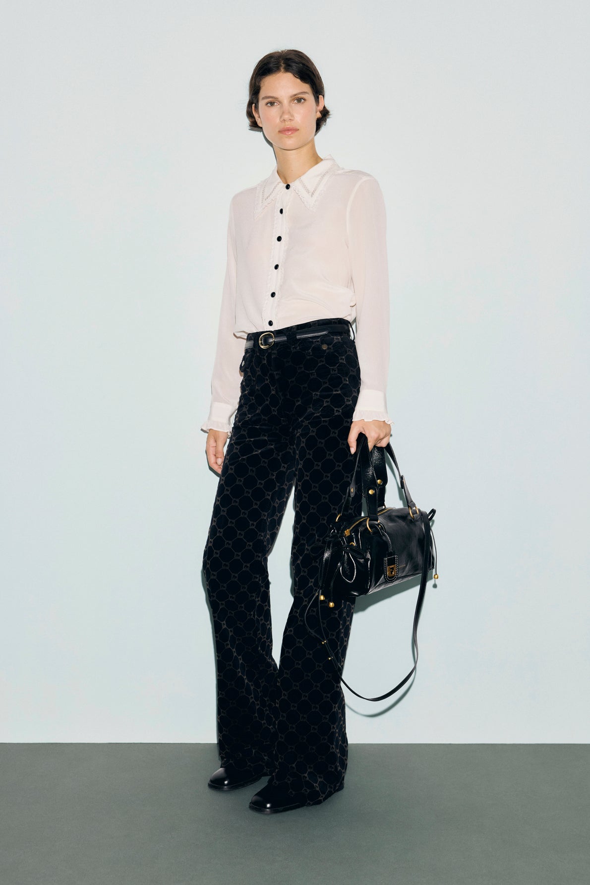 A person stands against a plain background wearing a white button-up shirt, black jean flare velours pants with taille haute and motif monogramme, and black shoes, holding a large black handbag.