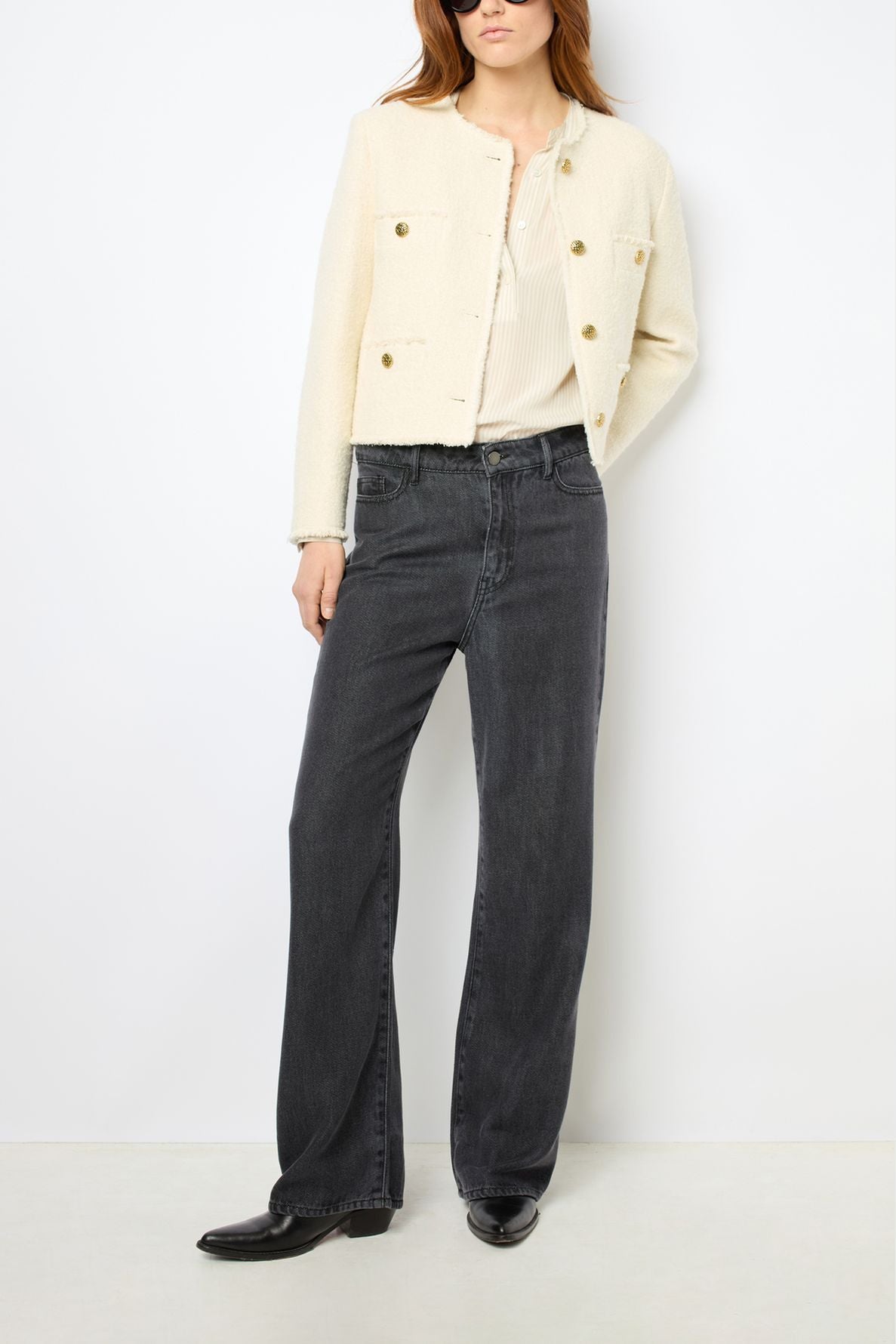 A person wearing a cream-colored buttoned jacket, a light top, dark wide-leg jeans, and black shoes stands against a white background.