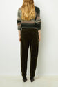 A person with long wavy hair, wearing a striped sweater, dark green pants, and black shoes, is standing facing a white wall with their back to the camera.