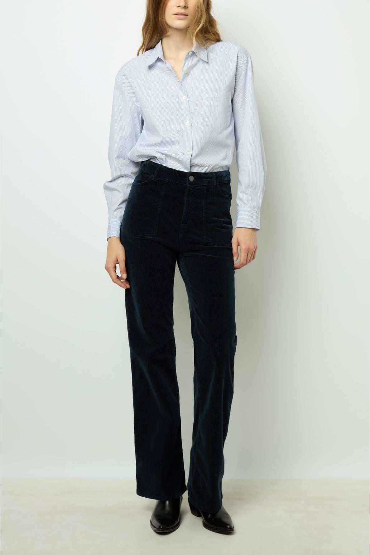 A person wearing a light blue button-up shirt, dark high-waisted pants, and black shoes stands against a plain white background. Their head is slightly tilted, and their left hand rests by their side.
