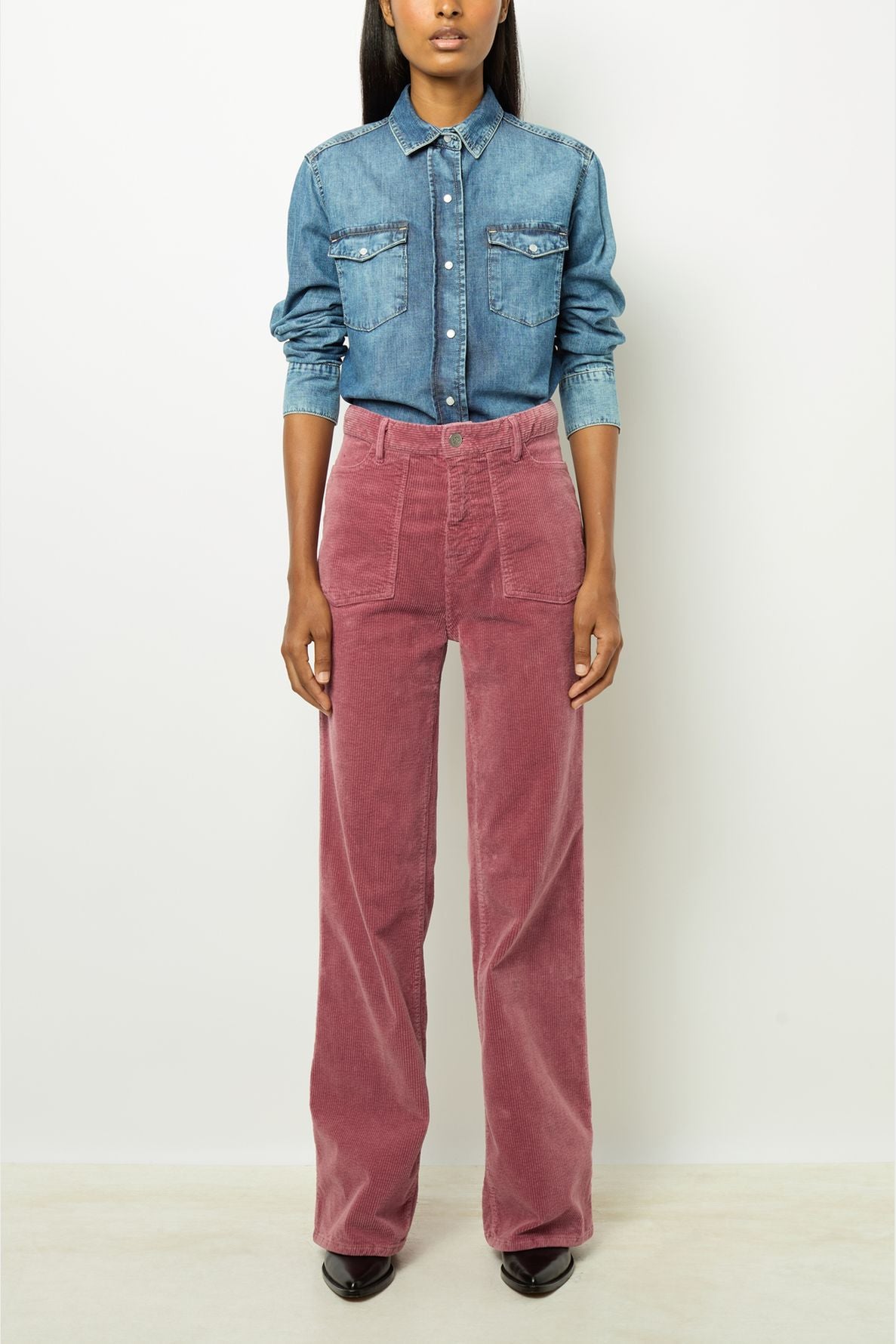 Person wearing a blue denim button-up shirt tucked into high-waisted, wide-leg pink corduroy pants, standing against a plain white background.