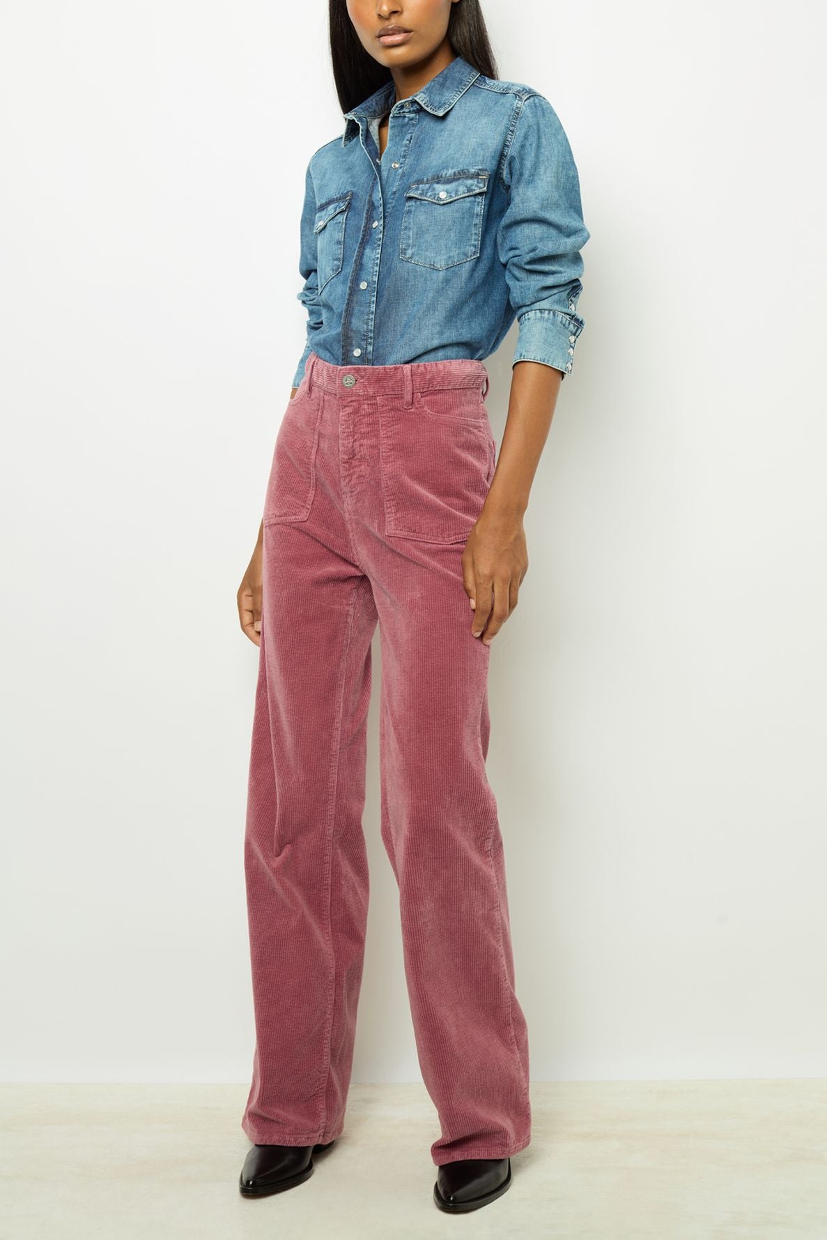 A person wearing a blue denim button-up shirt tucked into high-waisted, wide-leg pink corduroy pants with black shoes stands against a plain white background.