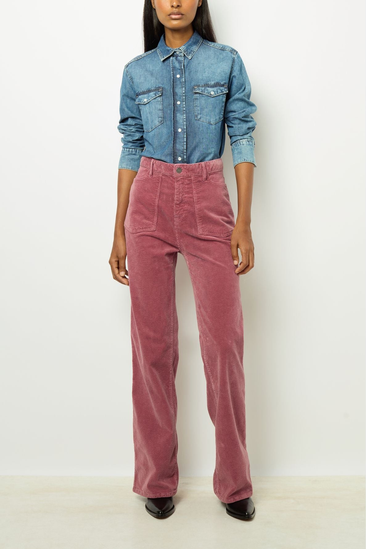 A person stands against a white wall wearing a blue denim button-up shirt with rolled sleeves and high-waisted pink corduroy pants, along with black pointed shoes.