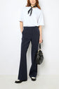 A person wearing a white t-shirt, dark pantalon large, black-and-white shoes, a black scarf, and holding a black handbag stands against a plain white background. The outfit features tissu uni for an effortlessly chic look.