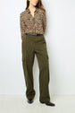Woman standing against a white wall, wearing a leopard print long-sleeve shirt tucked into high-waisted olive green cargo pants with a belt, and black shoes. Her arms are behind her back.