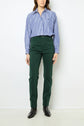 A person stands against a white wall wearing a blue and white striped button-up shirt, green pantalon coton coupe slim, and black ankle boots. The person’s hands are at their sides.