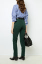 A woman stands facing away, wearing a blue striped shirt, dark green pantalon coton coupe slim, and black shoes, holding a black handbag in her left hand.
