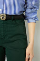 Person wearing a blue and white striped shirt, green pantalon coton with a coupe slim fit, and a black belt. Their sleeves are rolled up, and their right hand is visible with a small tattoo on the wrist. The image is cropped at the torso.