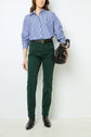 Person wearing a blue and white striped shirt, green pantalon coton, and black boots stands against a white background, holding a black handbag in one hand.