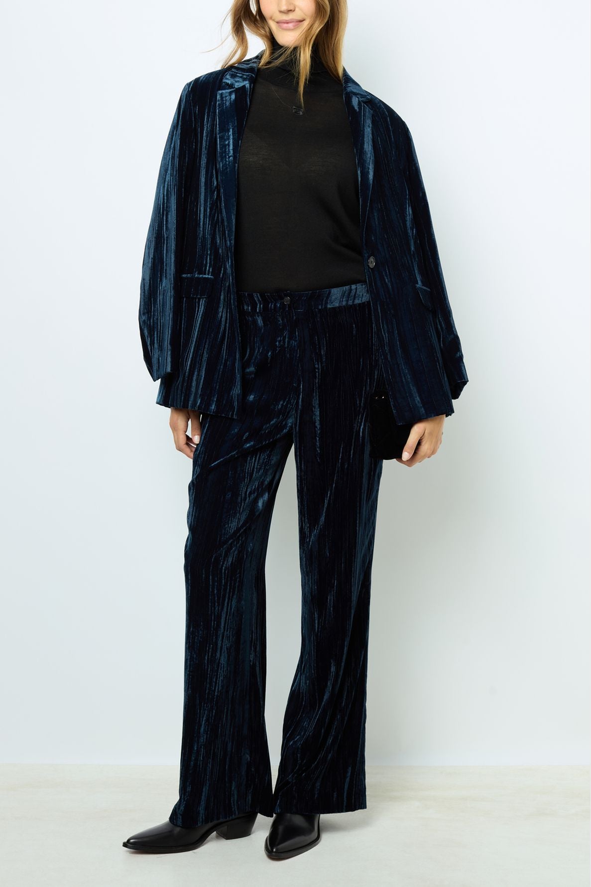 A woman wearing a dark velours frappé suit with matching jacket and pantalon long, paired with a black turtleneck and black pointed shoes, stands against a plain light background.