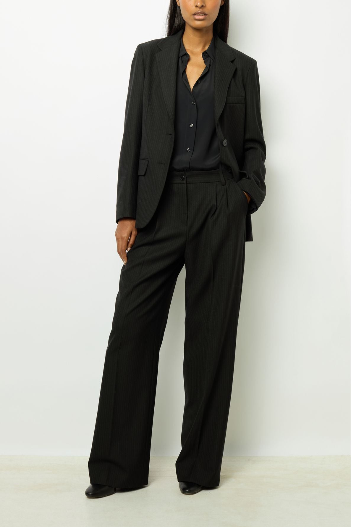 A person wearing a black suit with wide-leg trousers and a black shirt stands against a white background with one hand in their pocket.