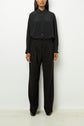 Person standing against a white wall wearing a loose black button-up shirt and high-waisted, wide-leg black trousers with black shoes. Their face is partially out of frame.