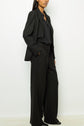 A person stands against a white background wearing a loose-fitting black suit with wide-leg trousers and a black shirt, hands in pockets. Their head is partly out of frame.