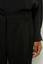 Close-up of a person wearing black pinstriped trousers and a black shirt. The persons right hand rests by their side, partially visible, and the shirt is tucked into the trousers.
