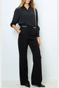 A woman stands against a white wall wearing a black button-up blouse, high-waisted black trousers with grommet details, black pointed shoes, and a crossbody bag. Her hands are in her pockets.