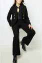 Woman sitting on a stool wearing a black textured suit with wide-leg pants, a black shirt, black boots, and a black necklace against a plain white background.