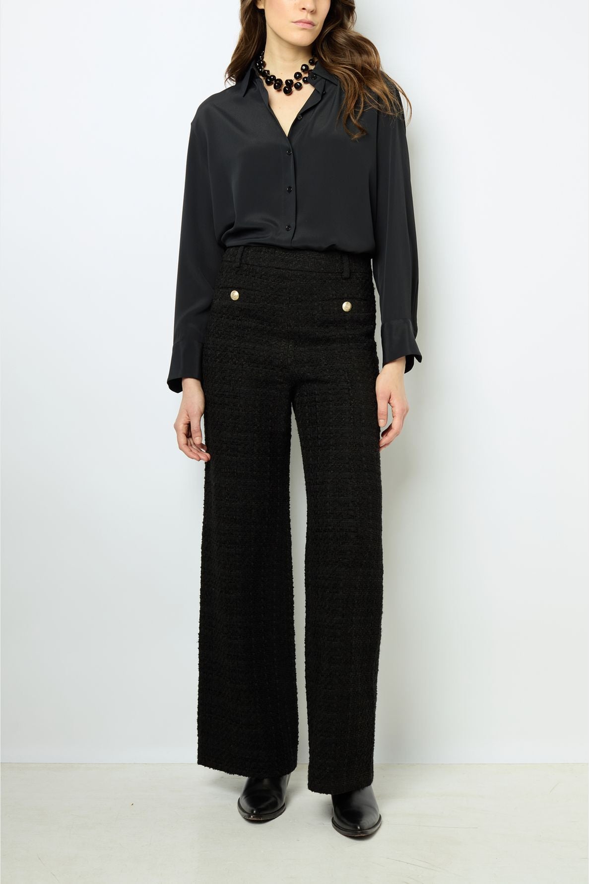 Woman wearing a black button-up blouse, high-waisted wide-leg textured black pants with buttons, black boots, and a chunky black necklace, standing against a plain white background.