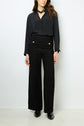 Woman wearing a black button-up blouse, high-waisted wide-leg textured black pants with buttons, black boots, and a chunky black necklace, standing against a plain white background.