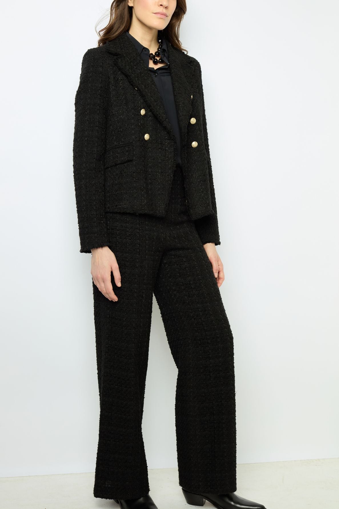 Woman wearing a textured black pantsuit with gold buttons, a black blouse, and black heeled boots, standing against a plain white background. Her face is partially out of frame.