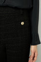 A person wears black textured pants with a gold button and a black long-sleeve blouse. Only the torso and right hand are visible against a plain background.