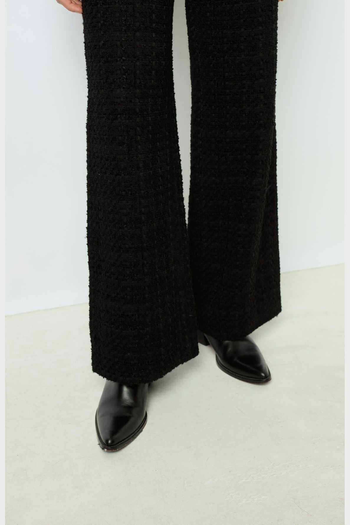 A person wearing textured black flared pants and black pointed-toe shoes stands on a light-colored floor against a plain white wall.