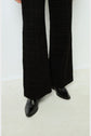 A person wearing textured black flared pants and black pointed-toe shoes stands on a light-colored floor against a plain white wall.