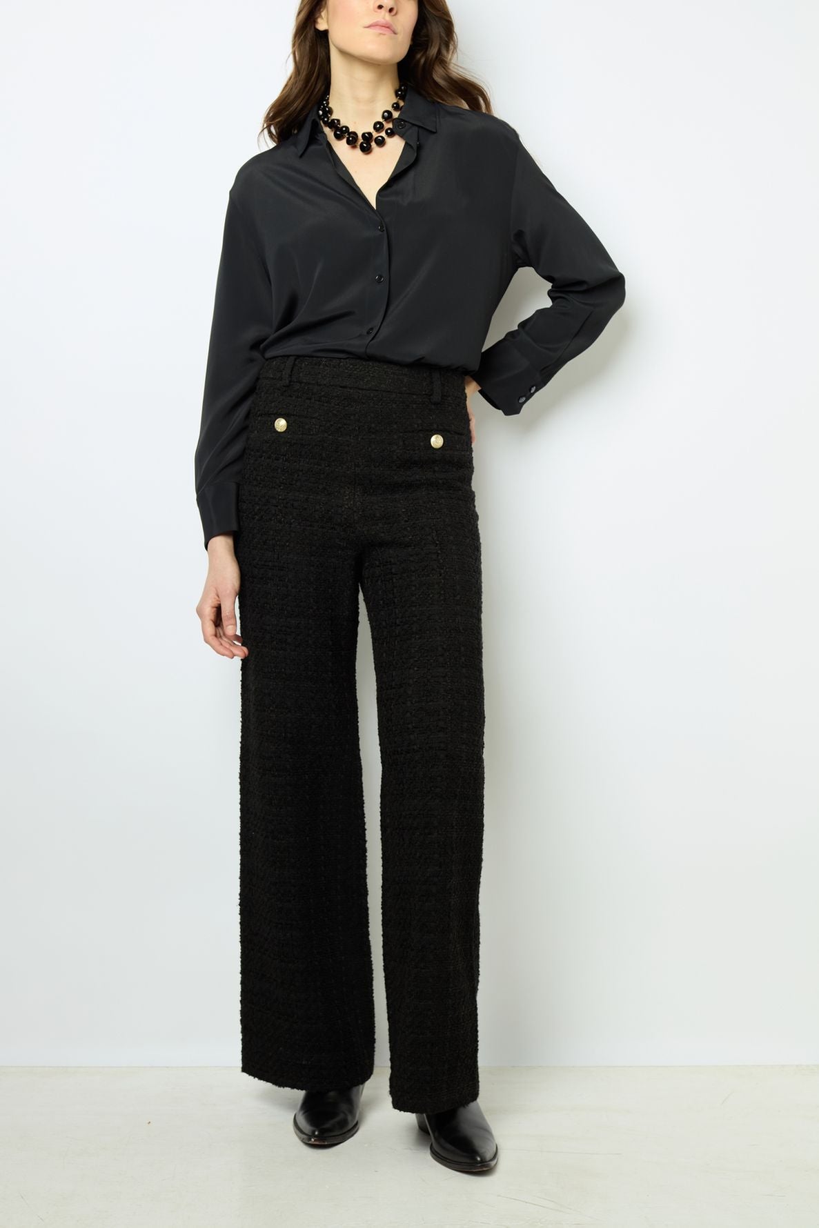Woman wearing a black button-up blouse, high-waisted black textured pants with gold buttons, black shoes, and a chunky black necklace, standing against a plain white background.