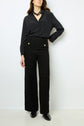 Woman wearing a black button-up blouse, high-waisted black textured pants with gold buttons, black shoes, and a chunky black necklace, standing against a plain white background.