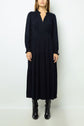 A person wearing a long-sleeved, ankle-length black dress with a v-neckline and black boots, standing against a plain light background.