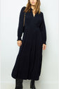 A woman wearing a long-sleeved, ankle-length black dress and black boots stands against a plain light background, with her left hand resting by her side.