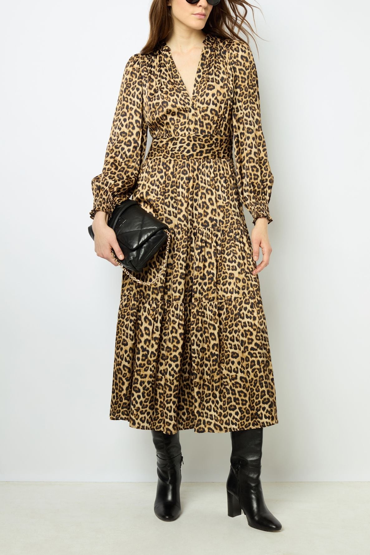 Woman wearing a long-sleeved, imprimé léopard robe midi satin, black knee-high boots, and sunglasses, holding a black quilted clutch handbag, standing against a plain light background.