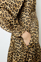 A person wearing a long-sleeved, glamour-chic robe midi satin with an imprimé léopard design, their right hand tucked into a side pocket. The focus is on the dress’s striking leopard print fabric and the elegant pocket detail.