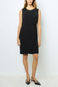 A woman wears a sleeveless black knee-length dress with a round neckline and black pointed-toe slingback flats, standing against a plain white background. This petite robe noire embodies effortless elegance, making it the perfect robe chic for any occasion.