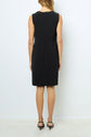 A woman stands facing away, wearing a chic petite robe noire— a sleeveless, knee-length black dress paired with black heels—against a plain white background.