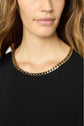 A woman wearing a black top reminiscent of a petite robe noire with a gold chain necklace, shown from the shoulders up. Her wavy light brown hair is visible, and only the lower part of her face is shown.
