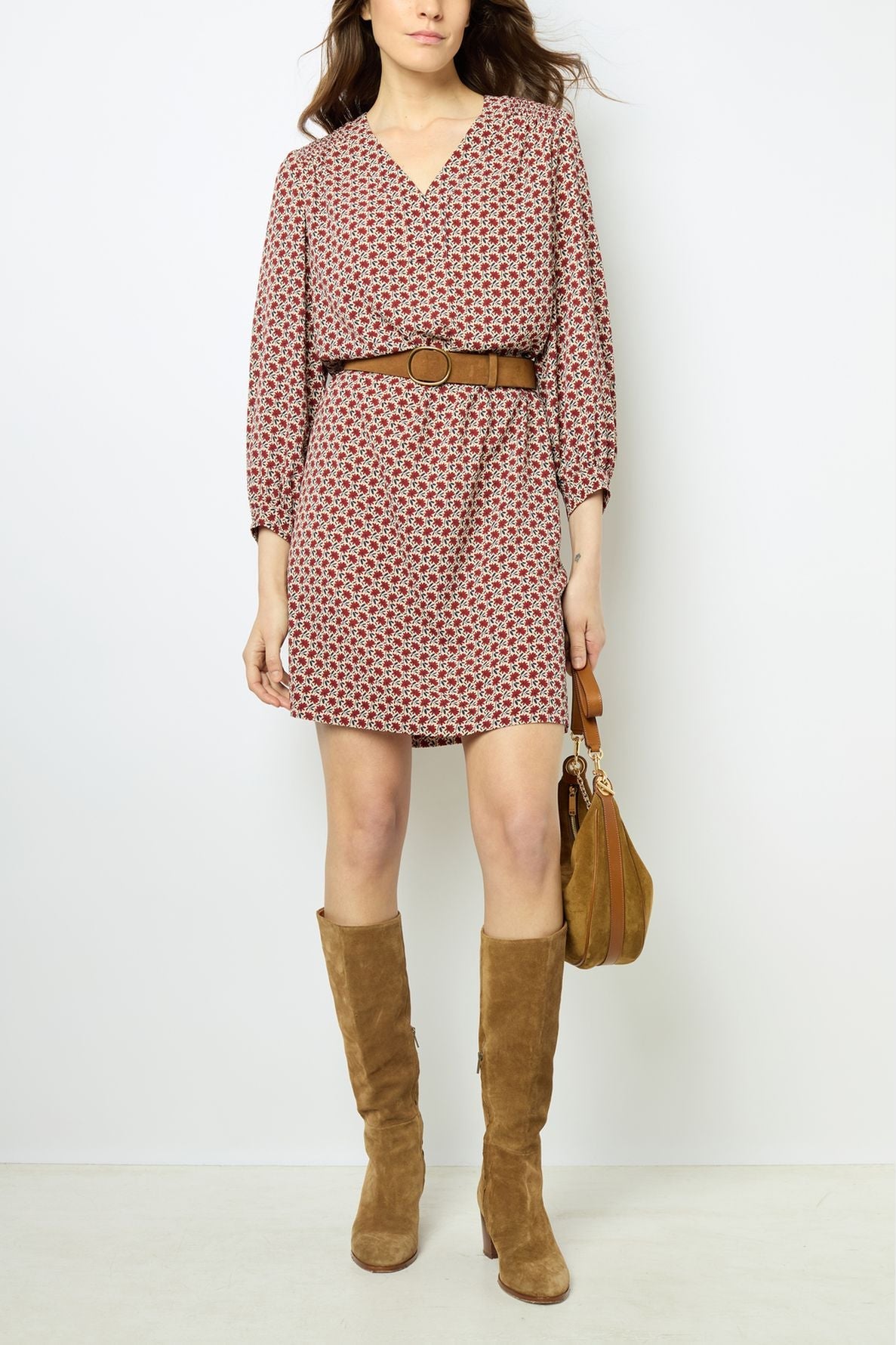 Woman wearing a motif floral robe courte with a brown belt, brown knee-high suede boots, and holding a brown handbag, standing against a plain white background.
