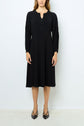 Woman standing against a white wall wearing a robe midi noire, long-sleeve and knee-length with a buttoned neckline and pleated details, paired with black low-heeled shoes.