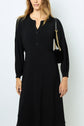 Woman wearing a robe midi noire à manches longues with a buttoned neckline and carrying a small black shoulder bag with a gold chain strap. Her hair is loose, and her face is partially out of frame.