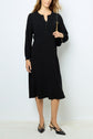A woman wearing a robe midi noire cintrée with buttons on the top, black slingback heels, and holding a small black shoulder bag, stands against a plain white wall.