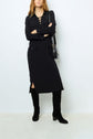 Woman wearing a black, long-sleeve, lace-up robe longue noire with front pockets, black knee-high boots, and a black shoulder bag, standing against a plain white background.